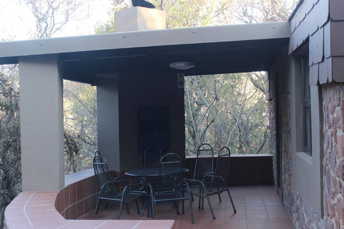 A covered patio area with a table and chairs, surrounded by stone walls and trees.