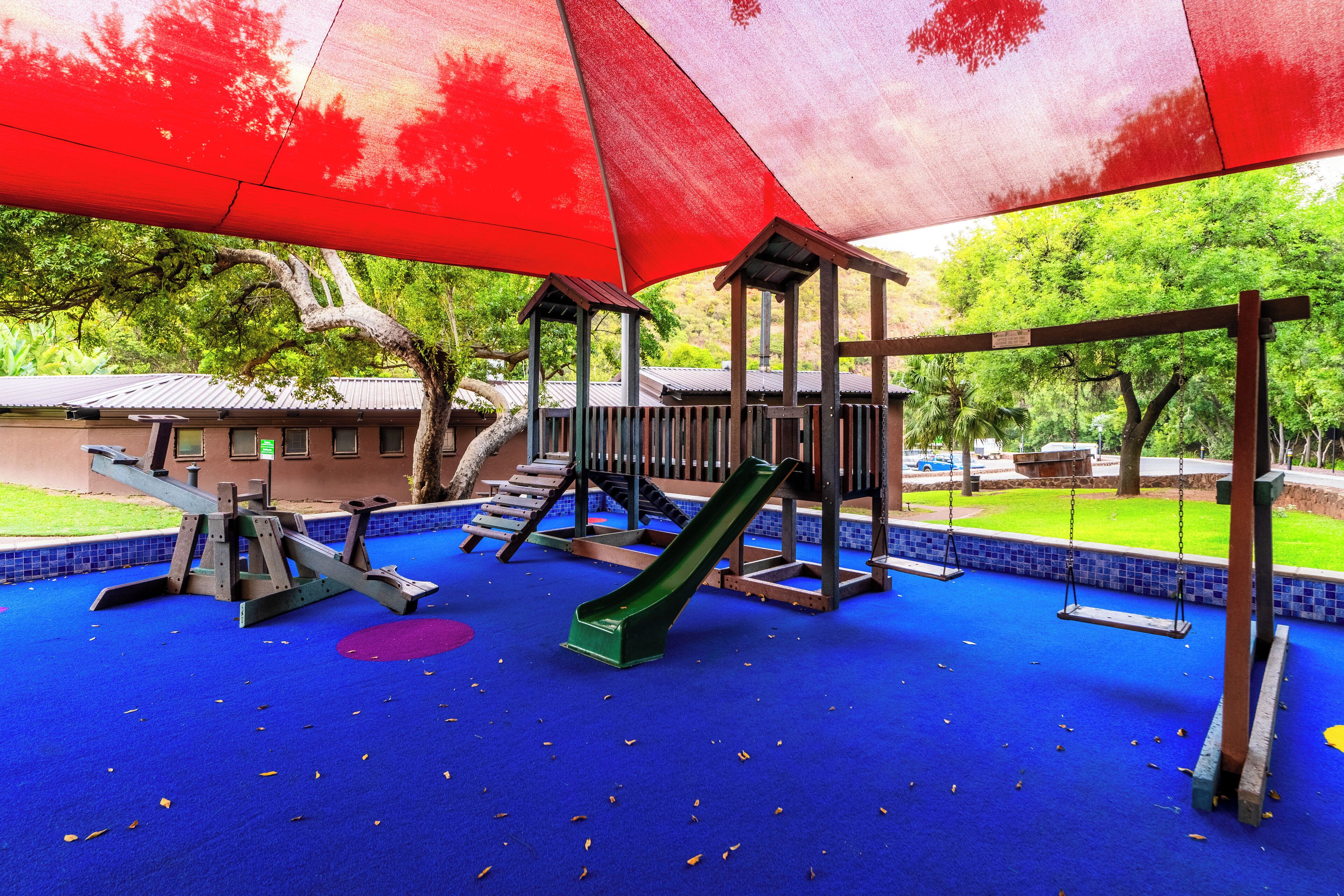A playground with a blue rubber surface, a green slide, and a swing set under a red canopy.