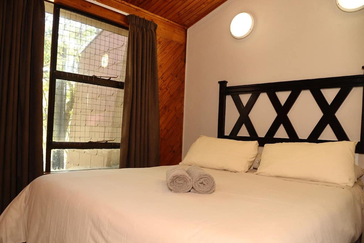 A bedroom with a wooden ceiling, a bed with white bedding, two pillows, and a window with curtains.
