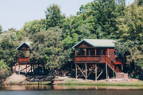 Deux cabanes en bois dotées de balcons, situées au bord d’un lac, entourées d’une forêt dense.