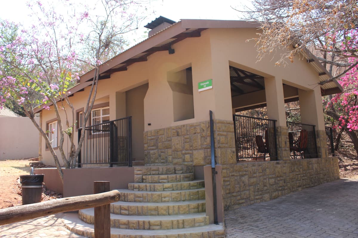 A building with a stone staircase leading to a covered entrance, a sign indicating the name, and a fenced area with chairs.