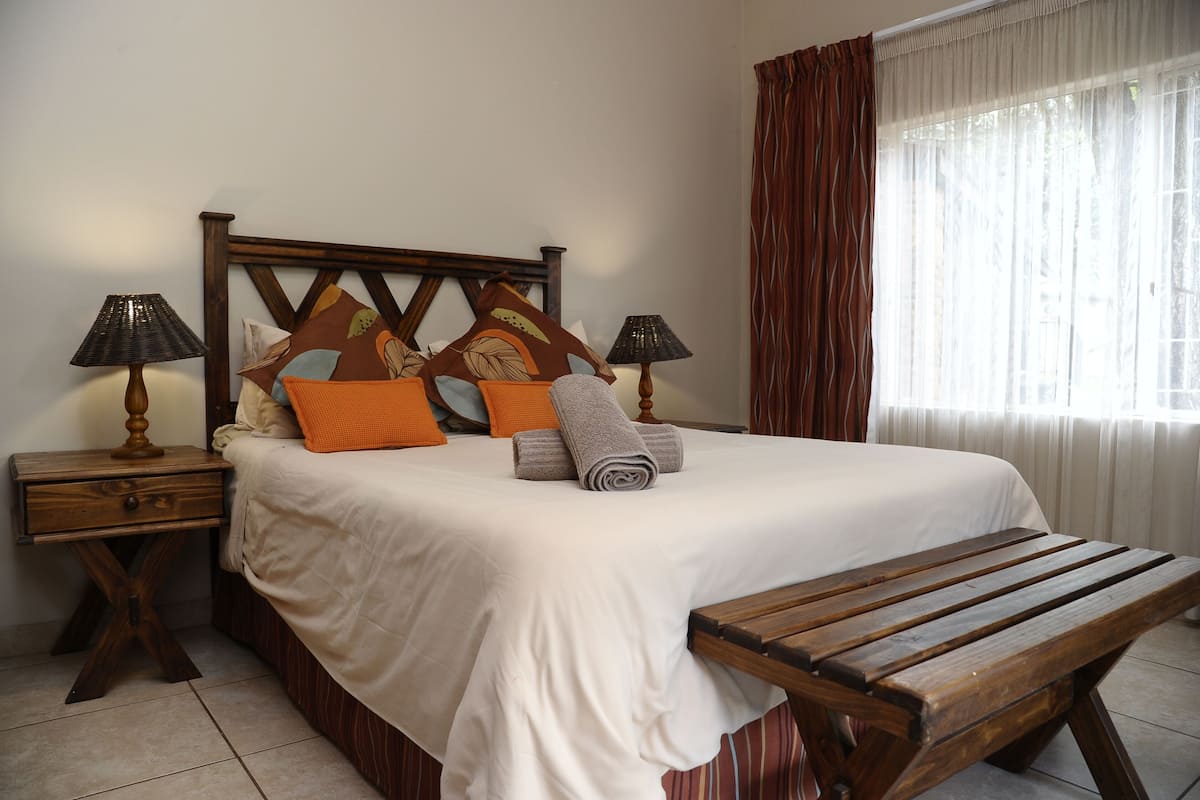 A neatly made bed with a wooden headboard, two bedside tables with lamps, a wooden bench, and a window with curtains.