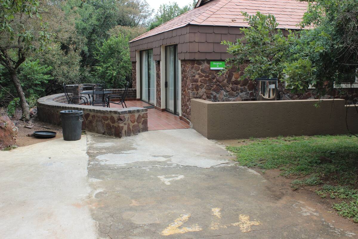A stone building with a tiled roof, a patio area with chairs and a table, and a trash can.
