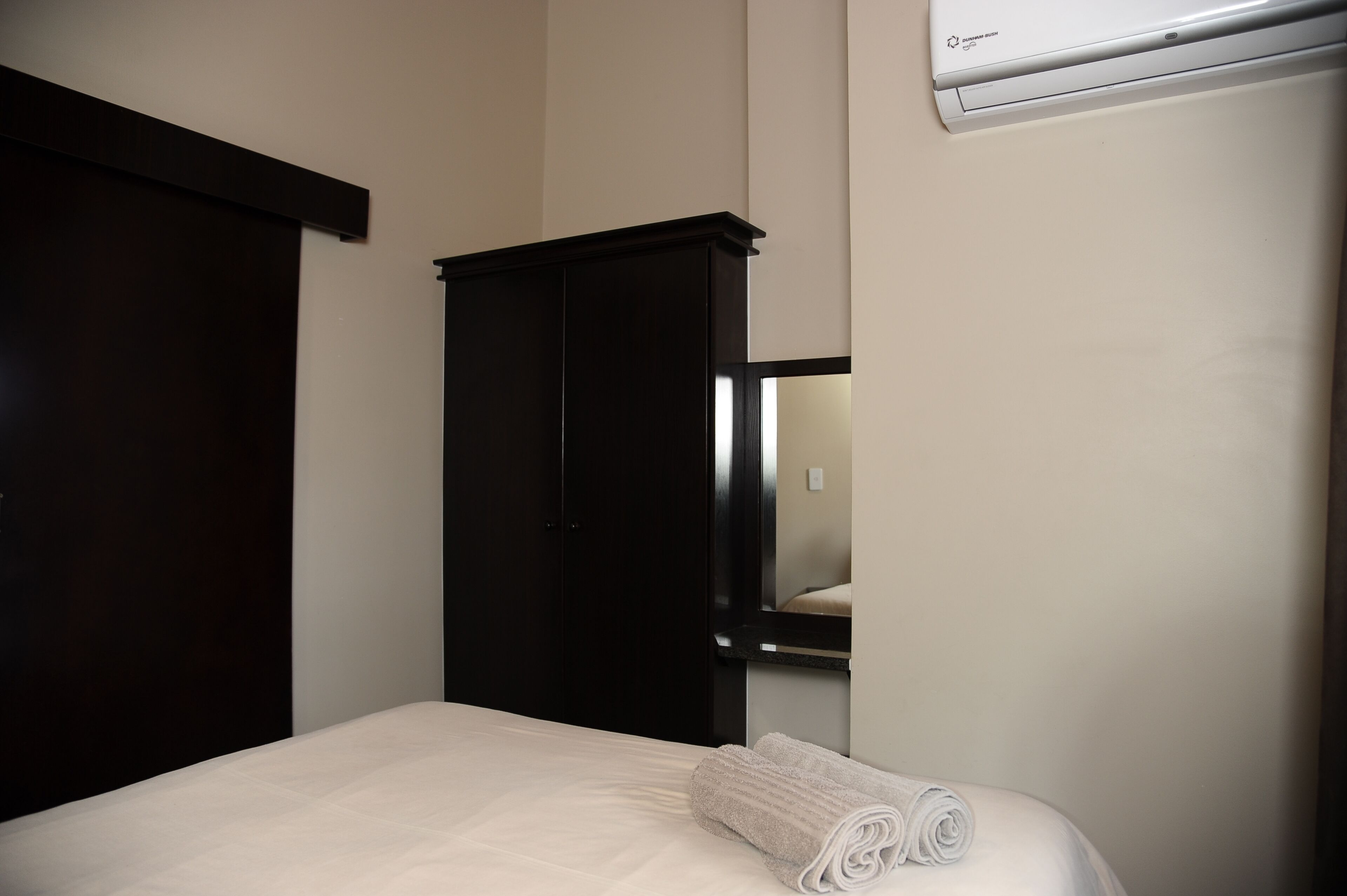 A hotel room with a bed, a dark wooden wardrobe, a mirror, and an air conditioning unit.