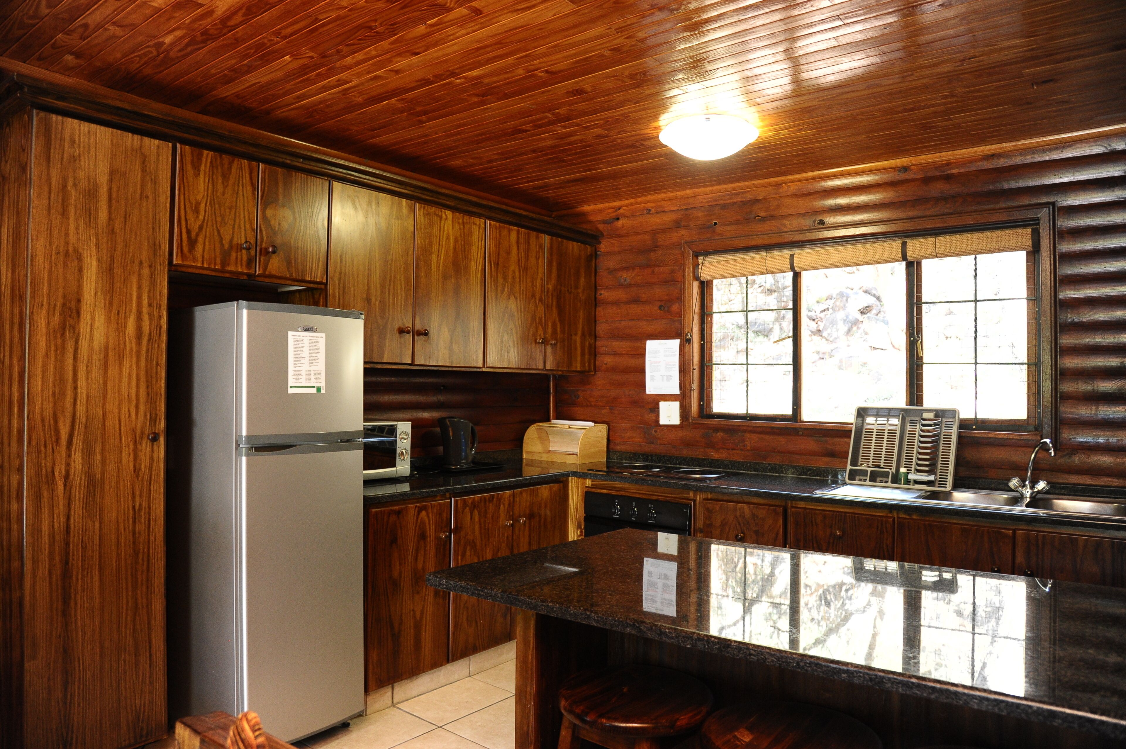 A kitchen with wooden cabinets, a refrigerator, a microwave, a toaster, and a window with blinds.