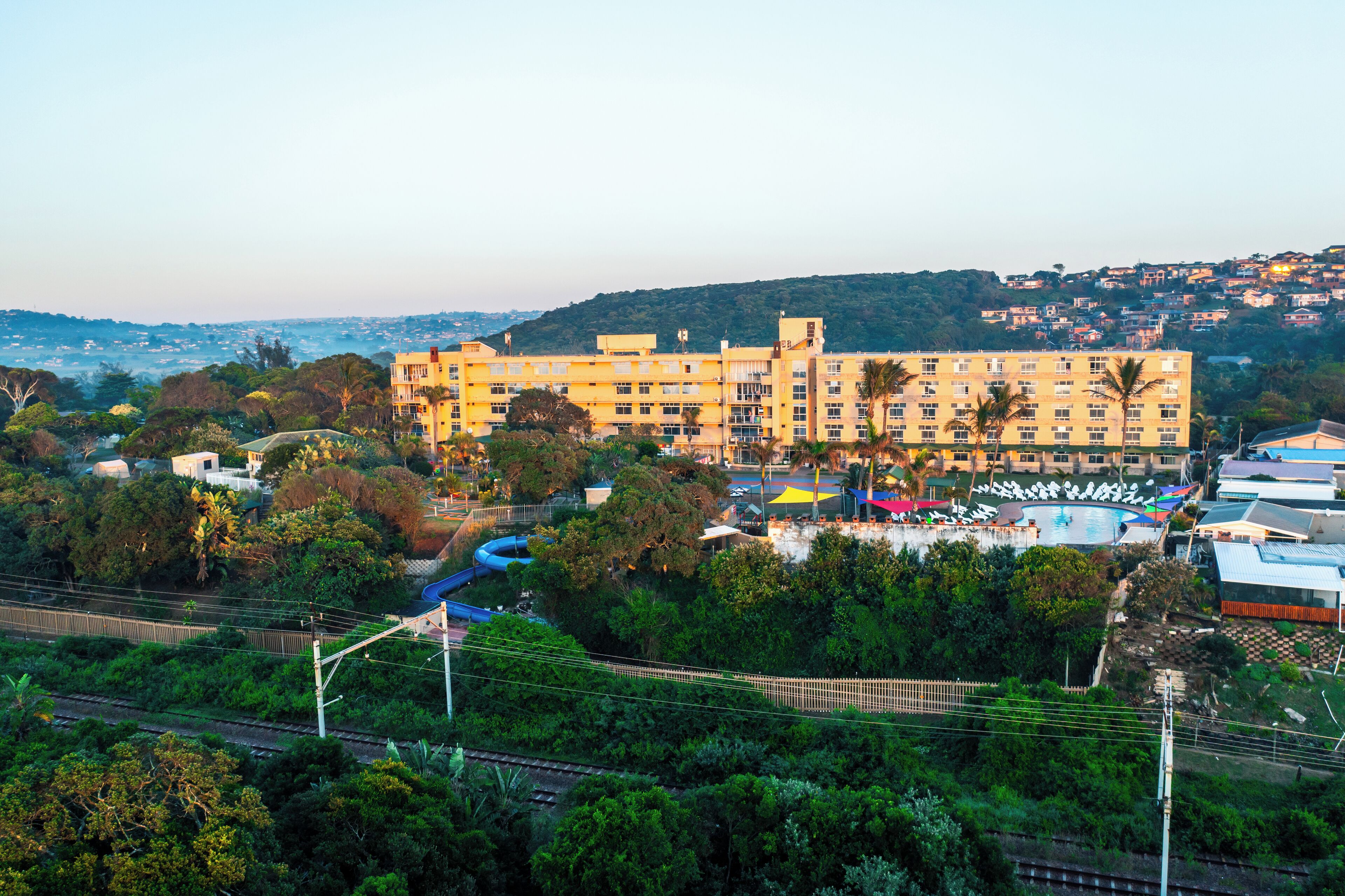 A large hotel with multiple floors, surrounded by lush greenery and a swimming pool.