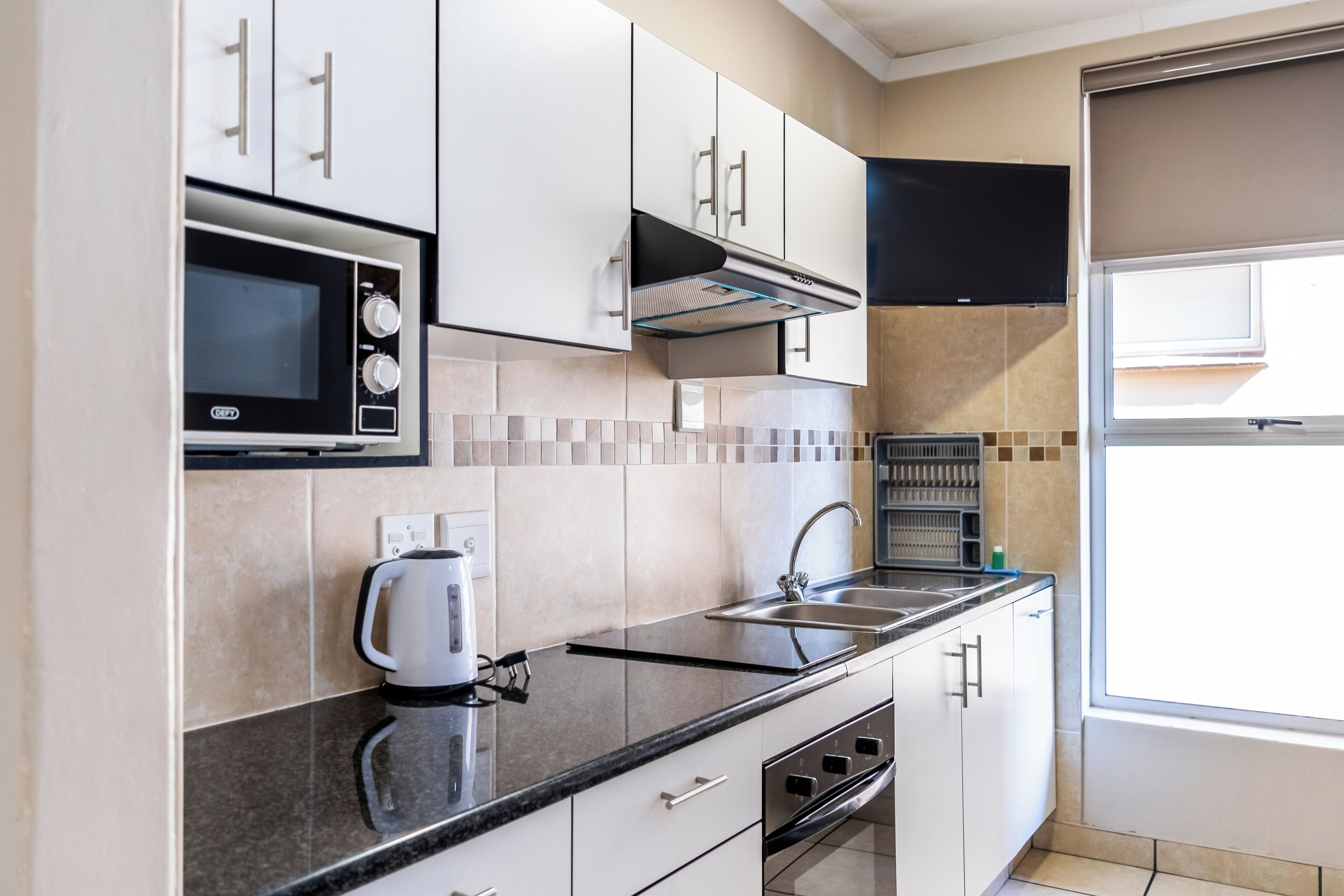 Una cocina moderna con armarios blancos, encimera negra, microondas integrado, hervidor y una ventana con persianas.