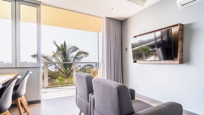 Ein modernes Wohnzimmer mit großem Fenster, grauem Sofa, Esstisch mit Stühlen und einem an der Wand befestigten Flachbildfernseher.