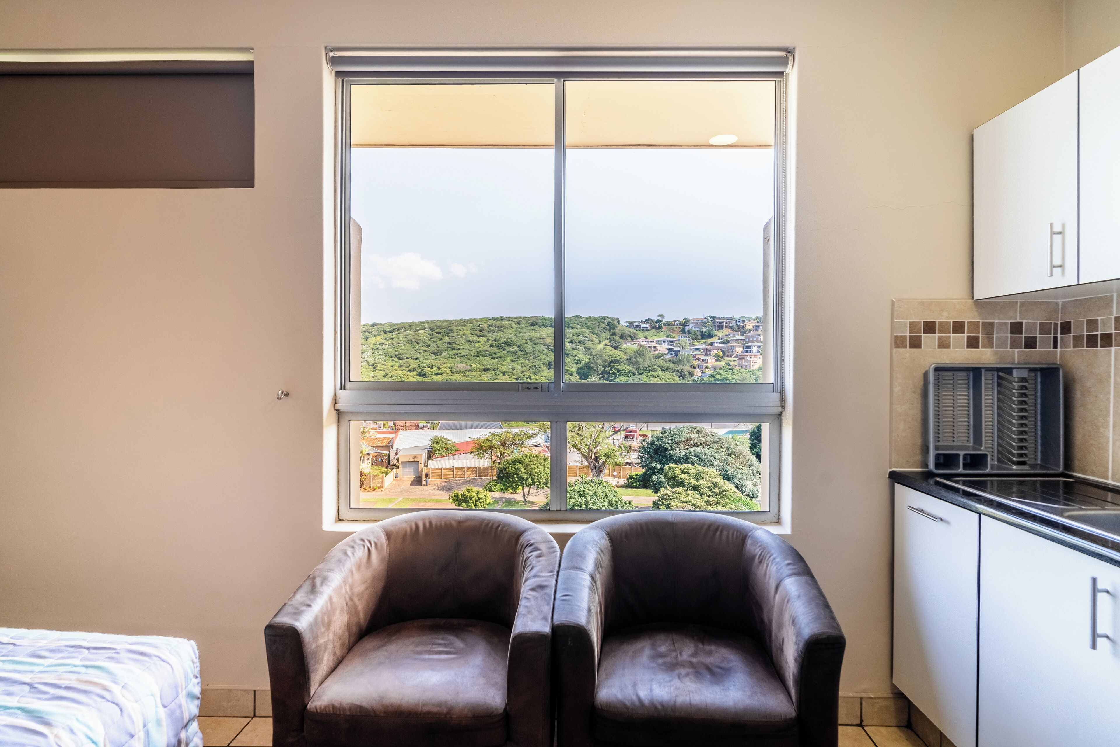 A room with a large window, two leather chairs, a bed, and a kitchenette.