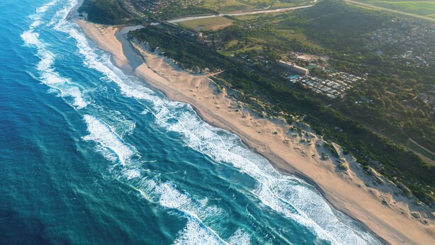 Luftaufnahme einer Küstenlandschaft mit Strand, Straße und verstreuten Gebäuden.