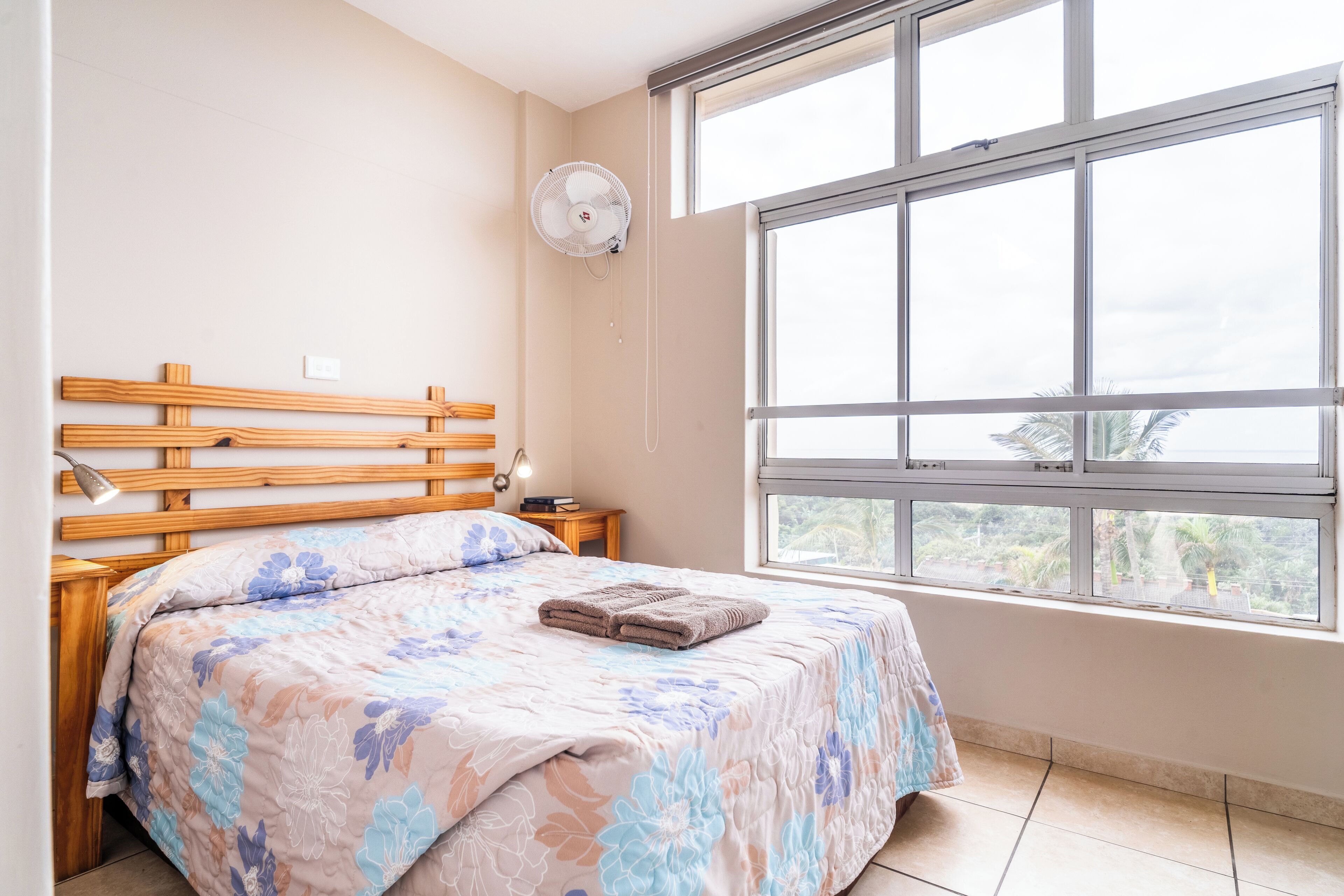 A bedroom with a wooden headboard, a large window with a view of greenery, a bed with floral bedding, and a bedside table with a lamp.