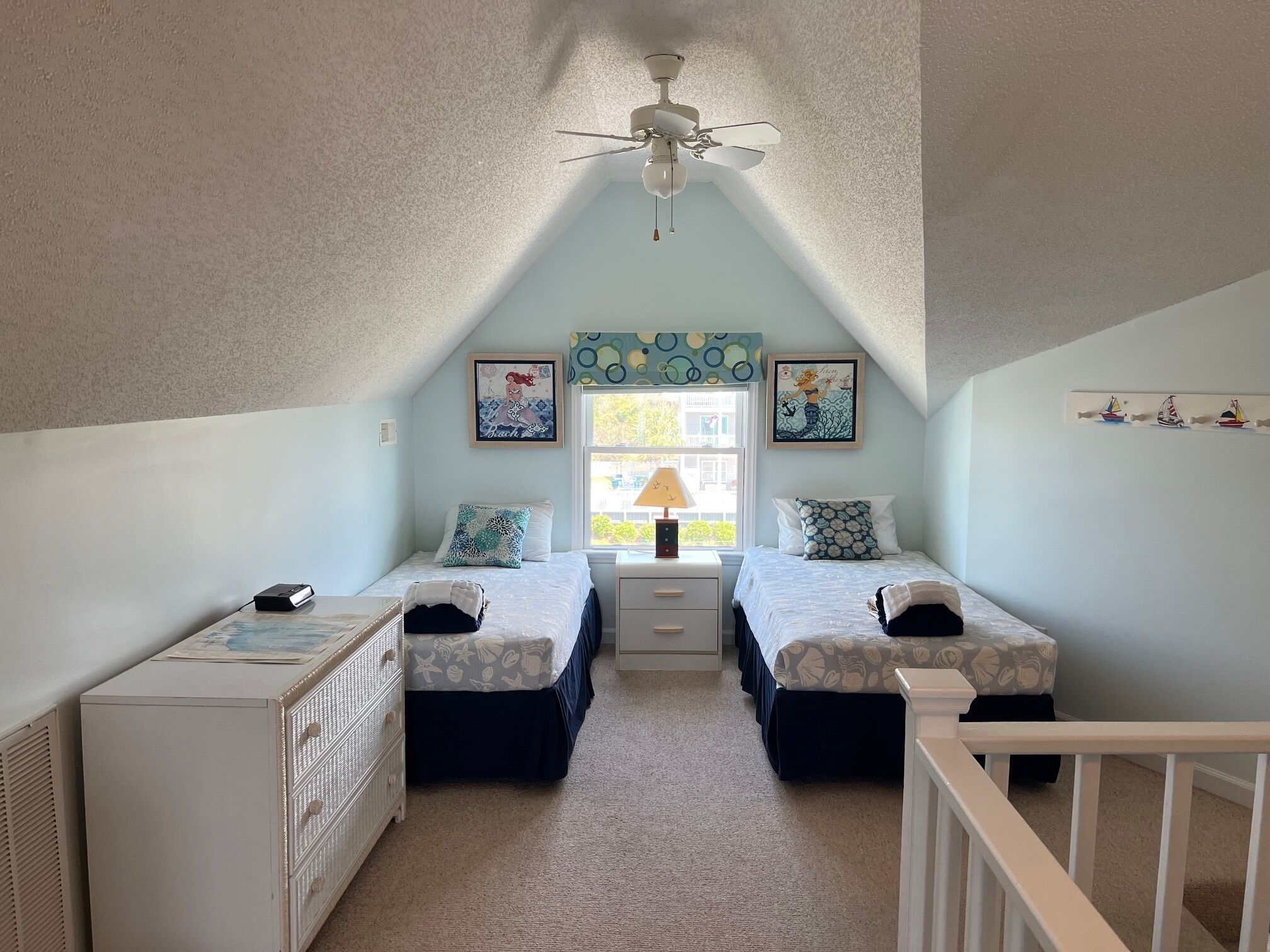 A bedroom with two single beds, a ceiling fan, a desk, and a window with a view of the outside.