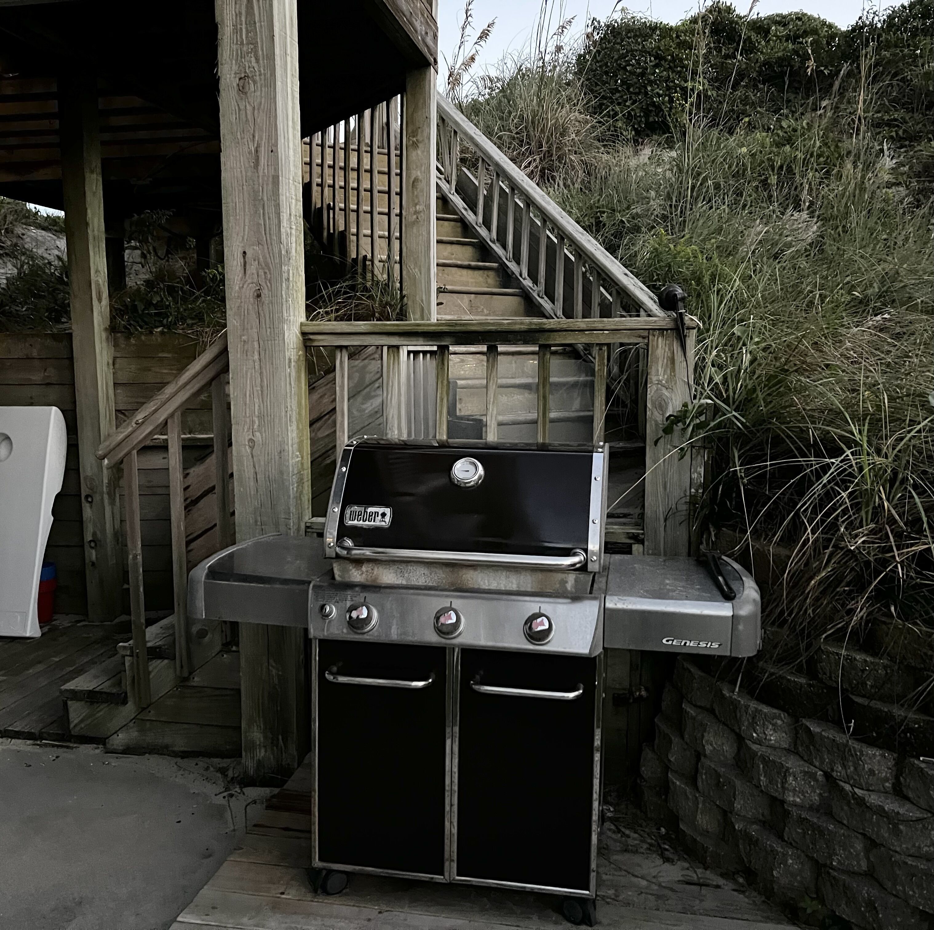 A Weber Genesis portable grill with a built-in oven, set up outdoors near a wooden staircase and a stone wall.