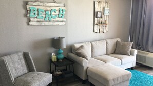 A living room with a grey armchair, a beige sofa, a teal rug, and wall decorations including a 'BEACH' sign and a 'Have a BODY 2 Go to the Beach' sign. - Lifes a beach on Myrtle Beach (Myrtle Beach)