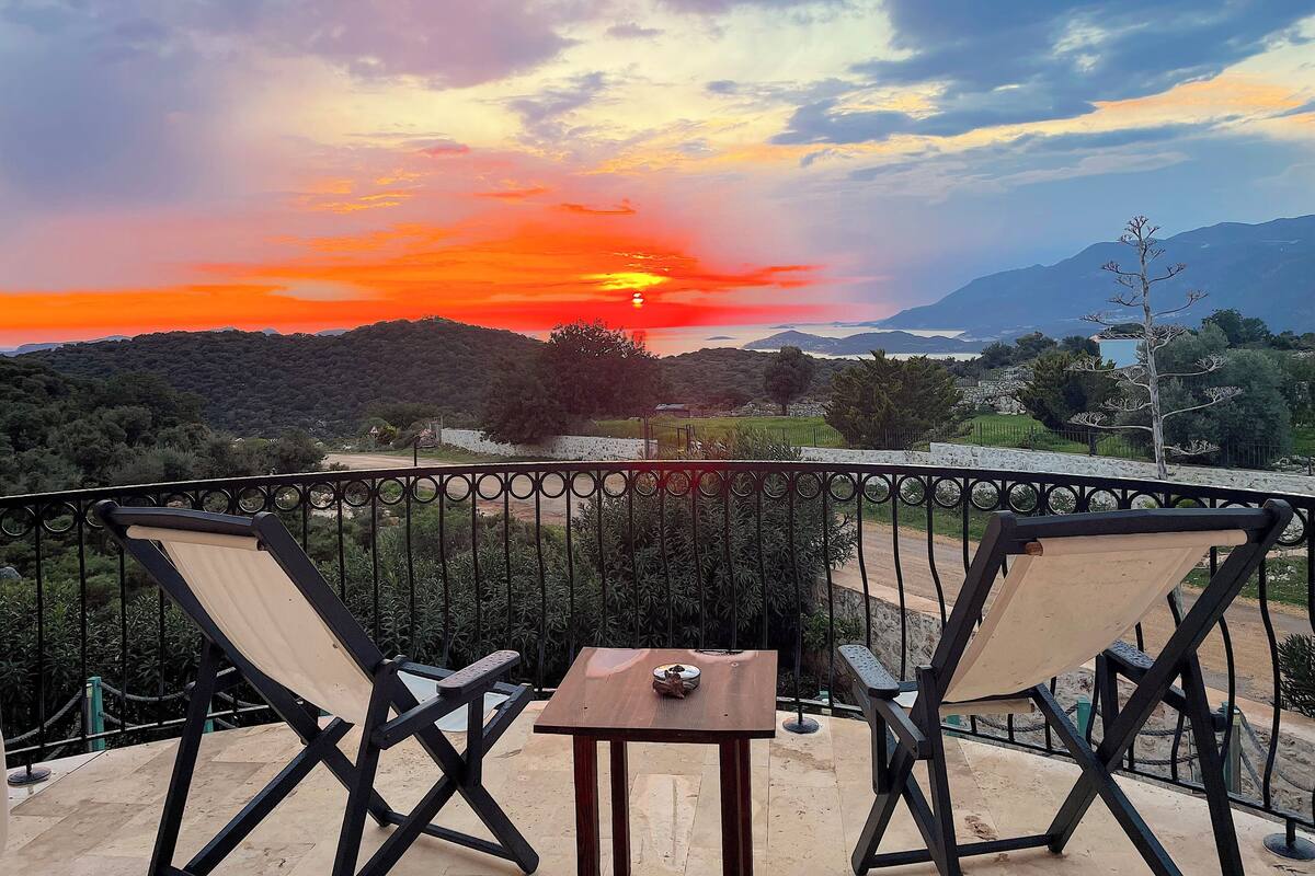 A balcony with two chairs and a small table, overlooking a scenic landscape with a sunset.