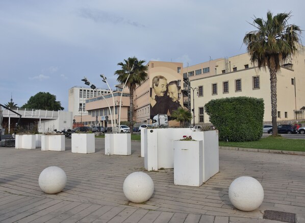A public square with white planters and spherical sculptures, a mural on a building, and palm trees. - Abside Suite & Spa (Palermo)