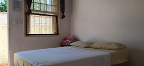 A small, simple room with a single bed, a window with a metal grill, and a wall-mounted light switch. - Pousada Recanto da Praia (Bertioga)