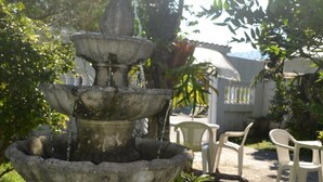 Una font de pedra de diversos nivells en un jardí, amb cadires i un edifici al fons.
