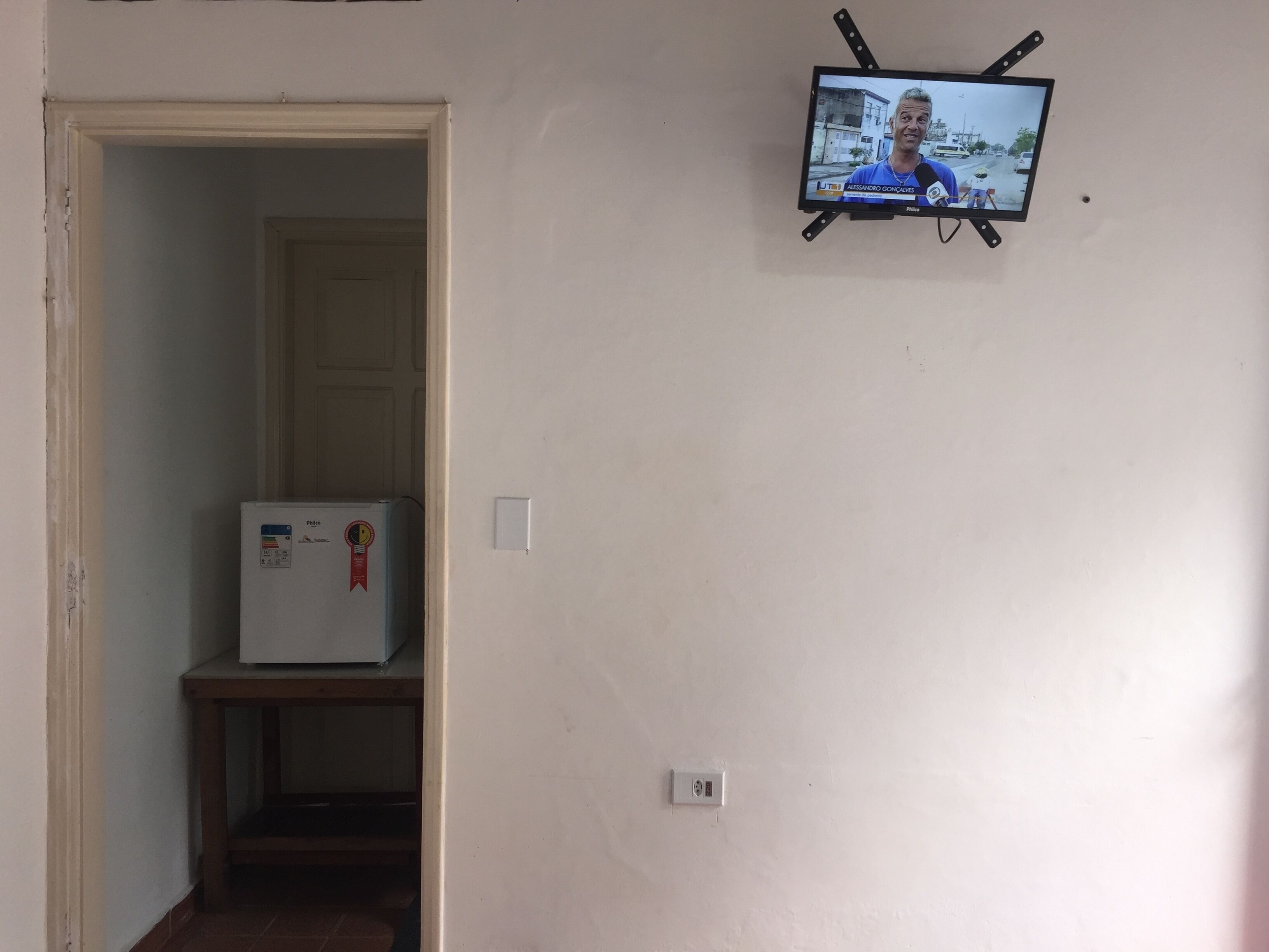 A hallway with a television mounted on the wall displaying a news program, a refrigerator, and a door leading to another room.