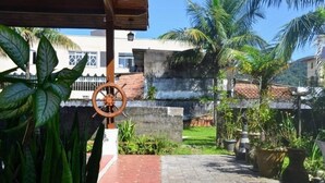 Un pati amb un cadireta de balanç de fusta, plantes en testos i vista a una zona residencial amb palmeres i un edifici blanc.