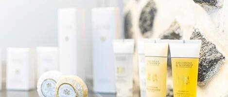 A selection of Gilchrist & Soames shampoo bottles and a bar of soap on a reflective surface, with a stone wall in the background.