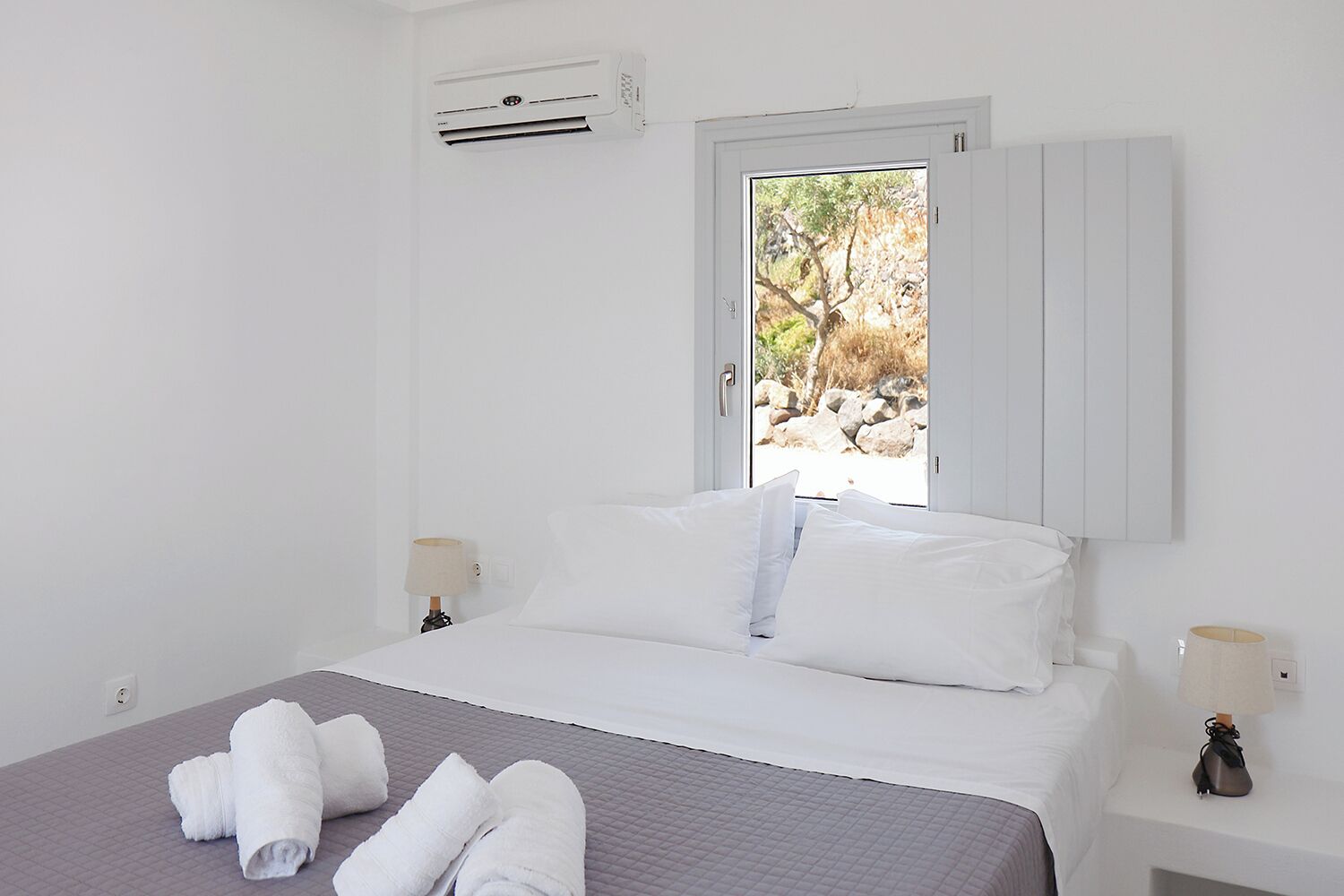 A hotel room with a bed, white walls, a window with a view of trees and rocks, an air conditioning unit, and two bedside lamps.