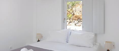 A hotel room with a bed, white walls, a window with a view of trees and rocks, an air conditioning unit, and two bedside lamps.