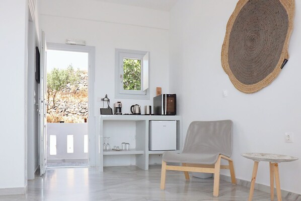 A modern, minimalist room with a white door leading to an outdoor area, a small kitchenette with a refrigerator and coffee maker, a grey chair, and a round woven wall hanging.