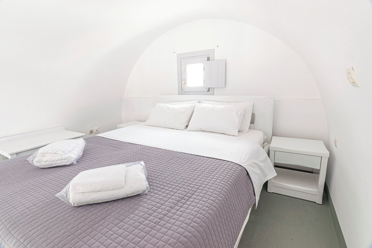 A bedroom with a bed, a nightstand, a bedside table, a window, and a wall-mounted light fixture.
