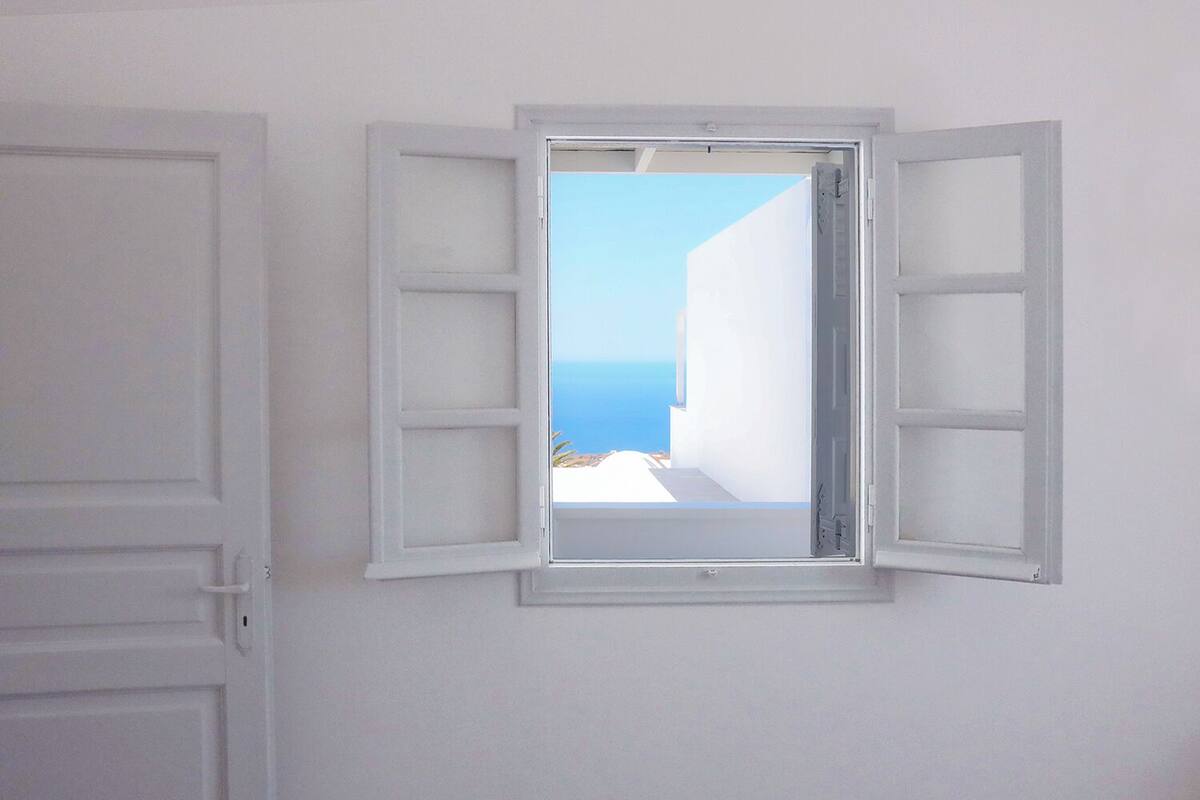 Ein Zimmer mit kleinem Fenster, Fensterläden, einer weißen Tür und Blick auf die Küste.