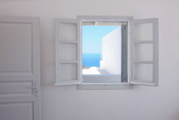 A room with a small window with shutters, a white door, and a view of a coastal area.