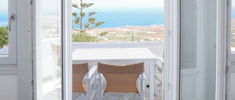 An open balcony with a white table and chairs, overlooking a coastal landscape with a tree and distant hills.
