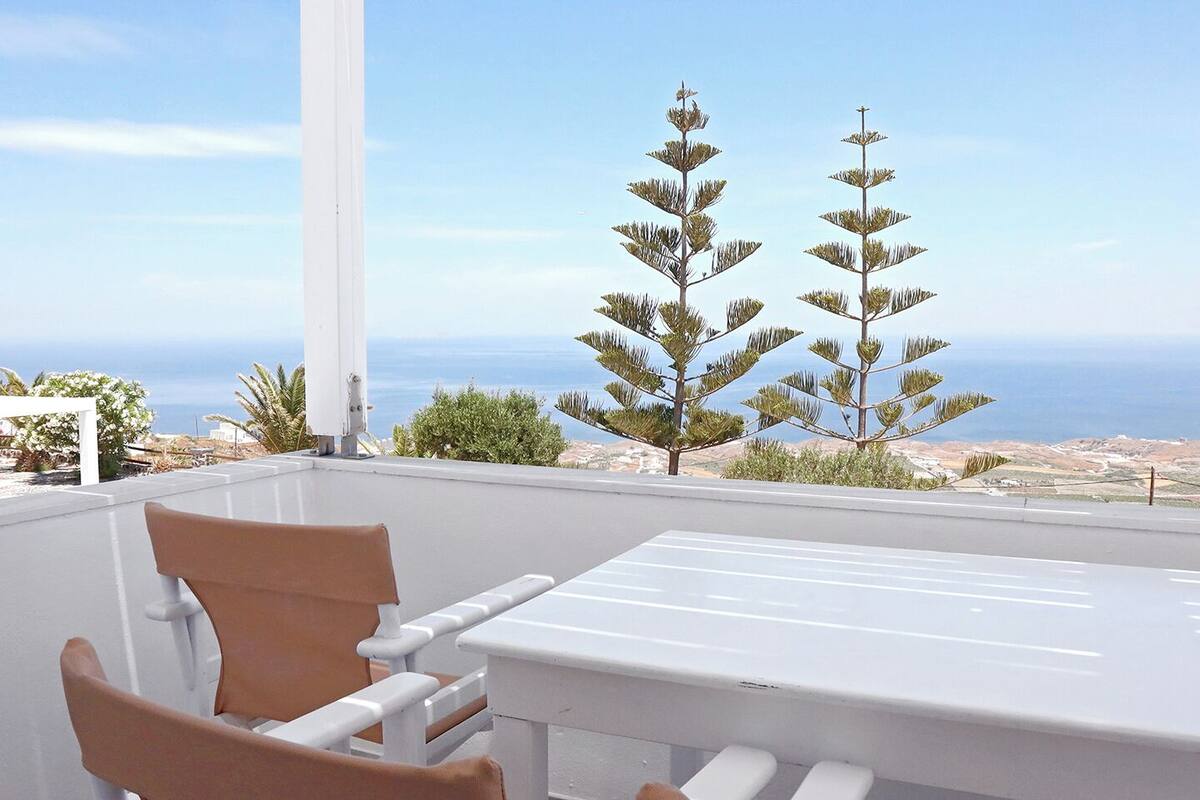 Eine Terrasse mit Tisch und Stühlen mit Blick auf eine Küstenlandschaft mit Bäumen und dem Meer.