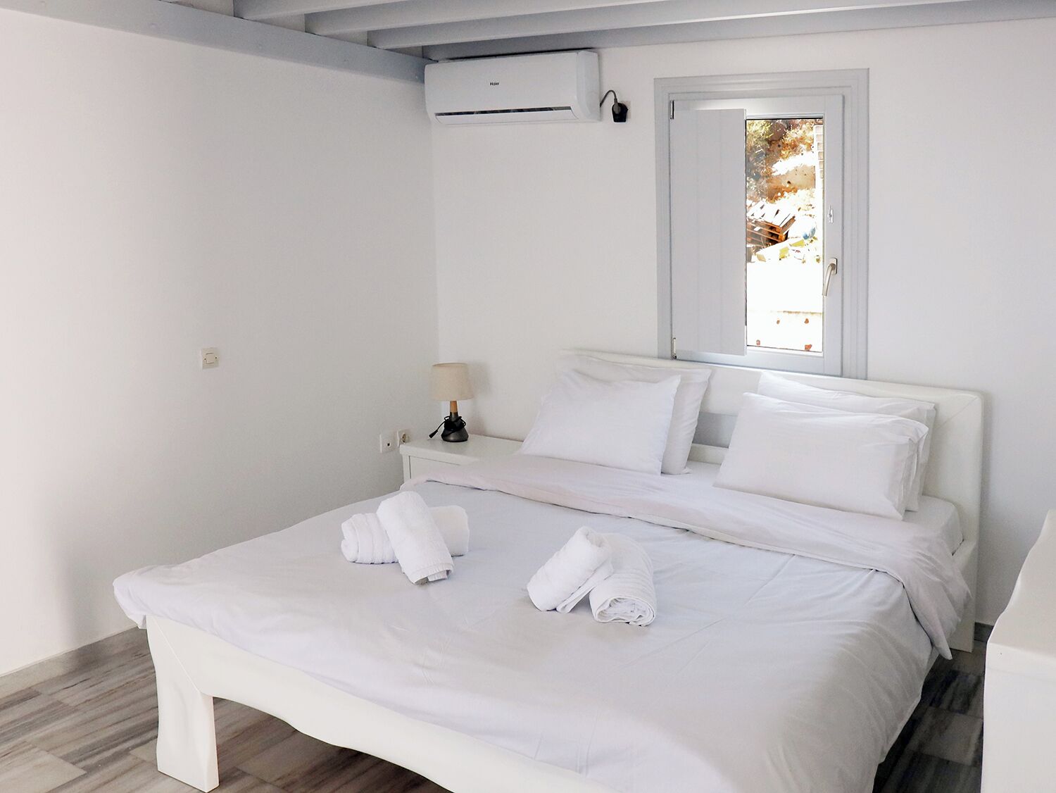 A clean, white hotel room with a bed, a bedside lamp, and an air conditioning unit.