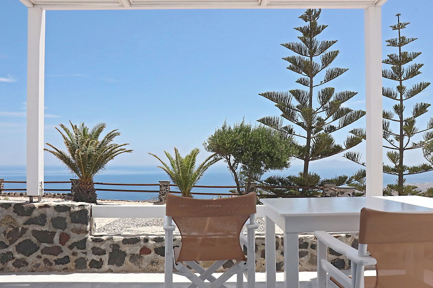 Una terraza con muros de piedra, una mesa blanca y sillas, con vistas al mar y árboles a la costa.