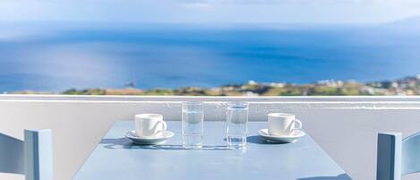 A table with two white cups and two glasses of water on a blue table, overlooking a coastal view.