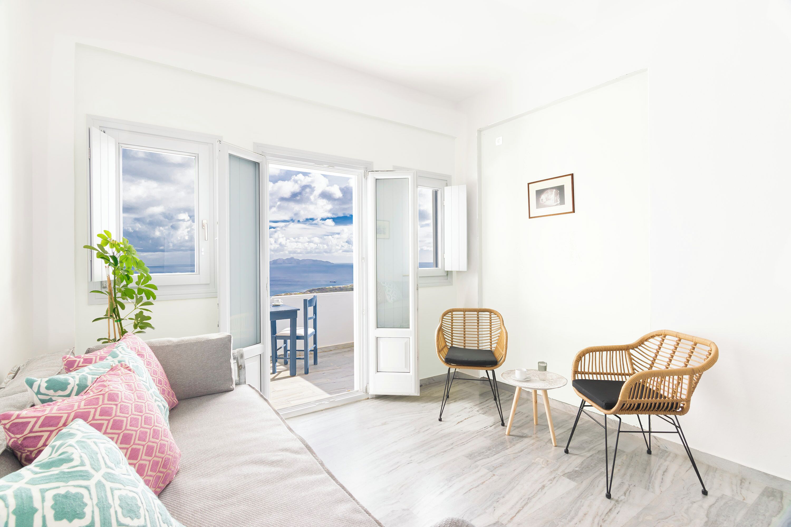 En lys stue med en sofa, to fletstole, et lille bord og udsigt til en balkon gennem åbne døre.