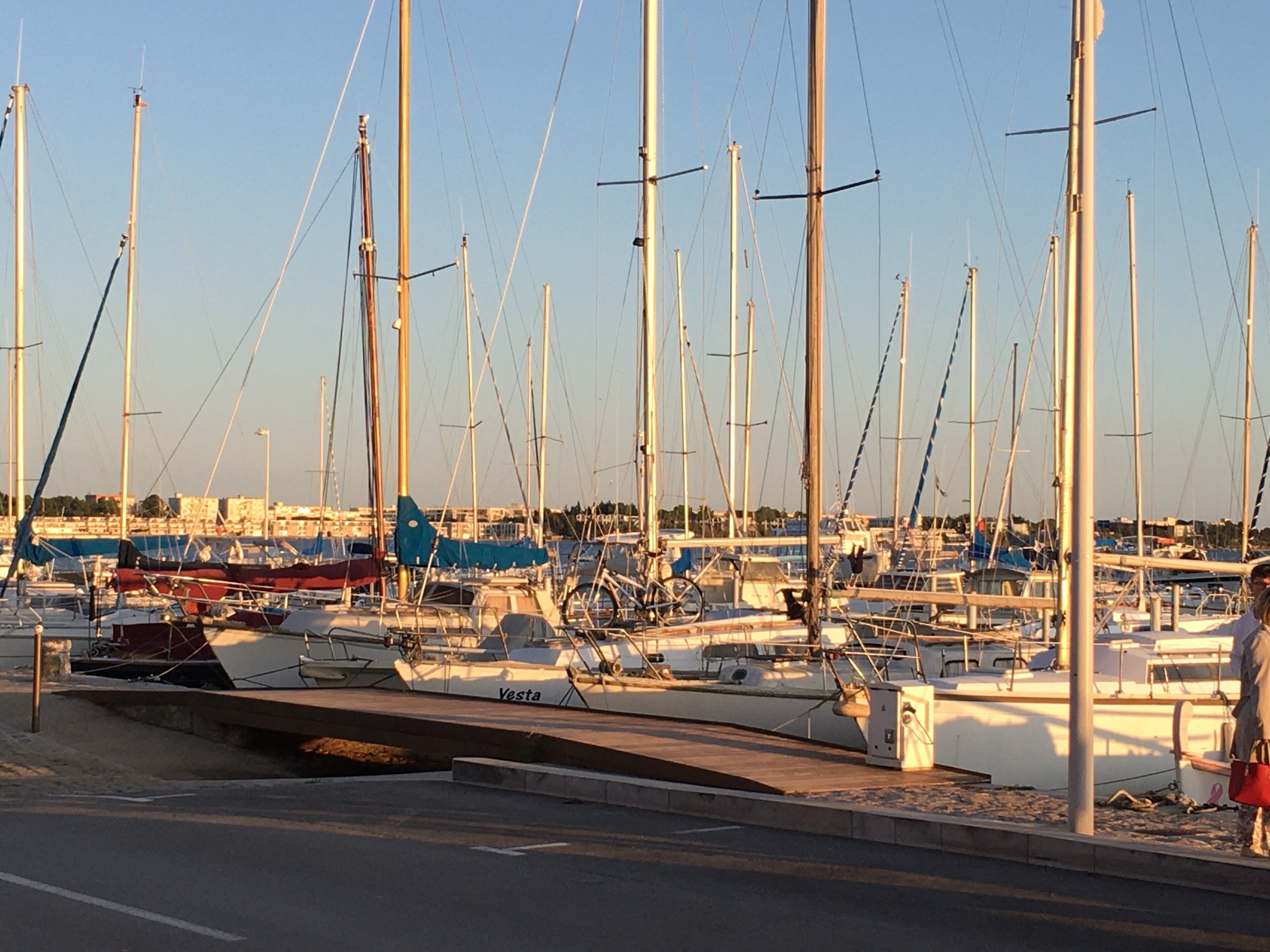 Un puerto deportivo con varias embarcaciones amarradas, un paseo marítimo y un cielo despejado.