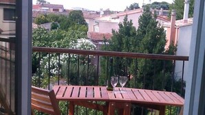 Une terrasse en bois avec une table et des chaises, deux verres à vin posés sur la table, et une vue sur des bâtiments et des arbres en arrière-plan.