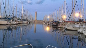 Un port de plaisance où sont amarrés de nombreux bateaux, un phare en arrière-plan et une eau calme qui reflète les bateaux et le ciel.