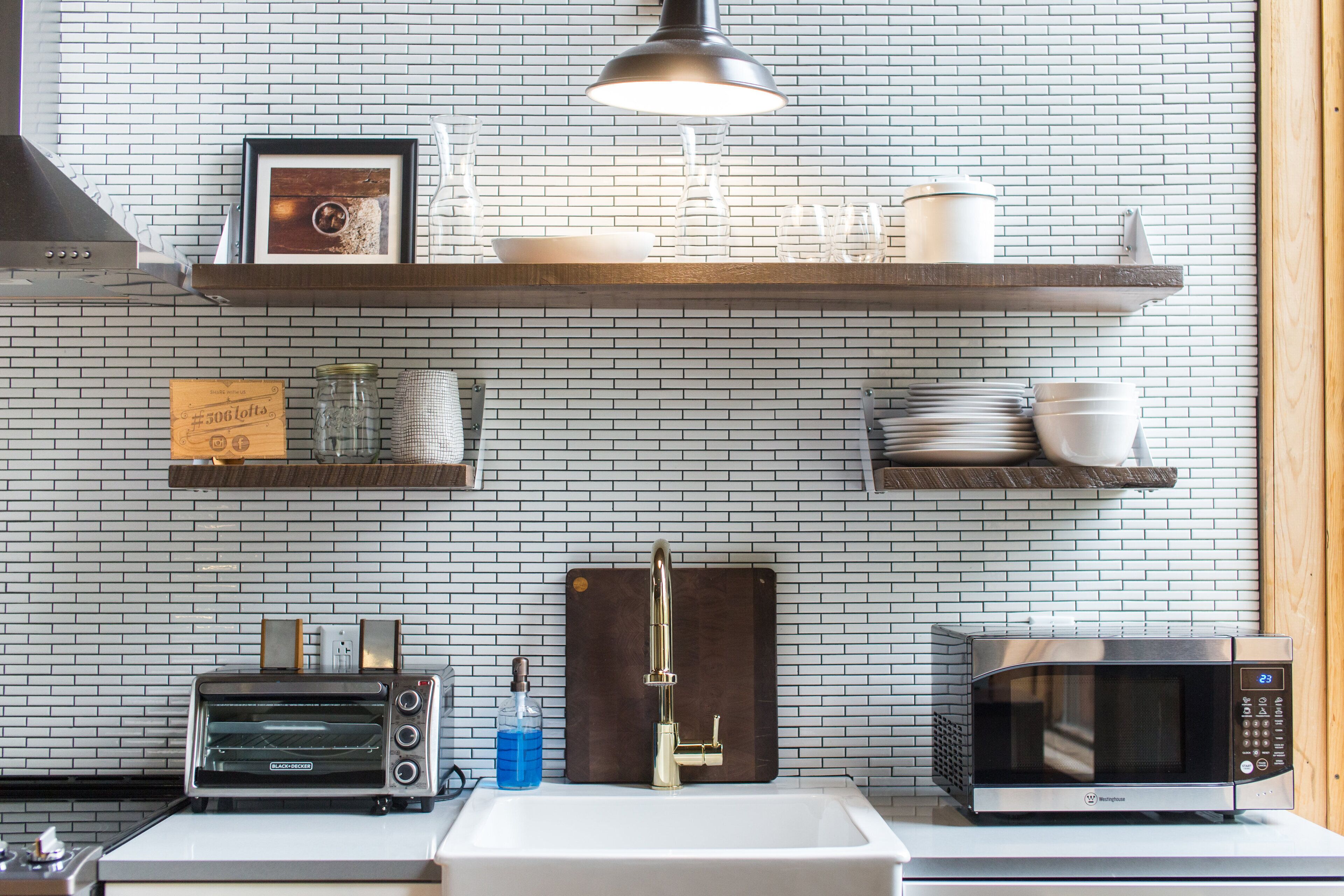 A kitchen with a white brick backsplash, a wooden shelf with dishes, a toaster, a sink, and a microwave.