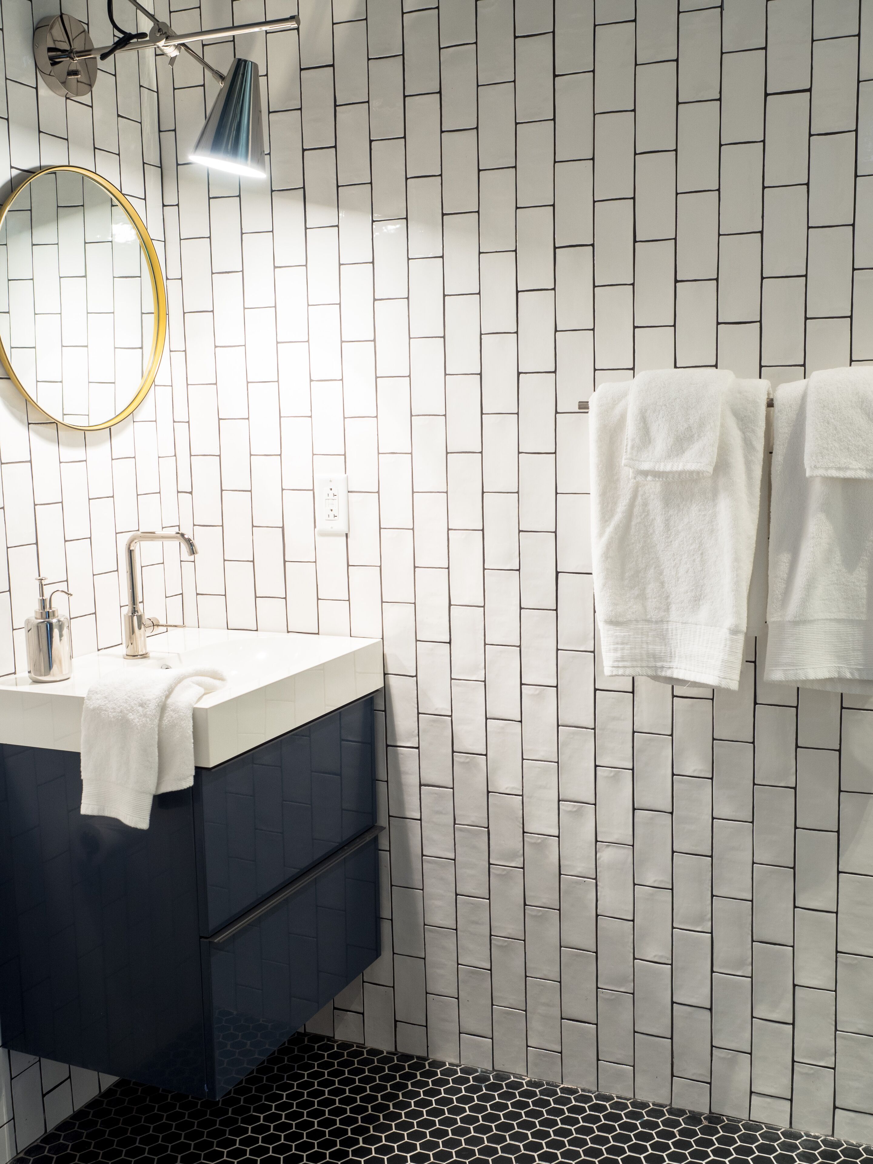 A bathroom with a white tiled wall, a dark blue vanity, a round mirror with a gold frame, a white sink, a faucet, a towel rack with two white towels, and a black and white patterned floor.