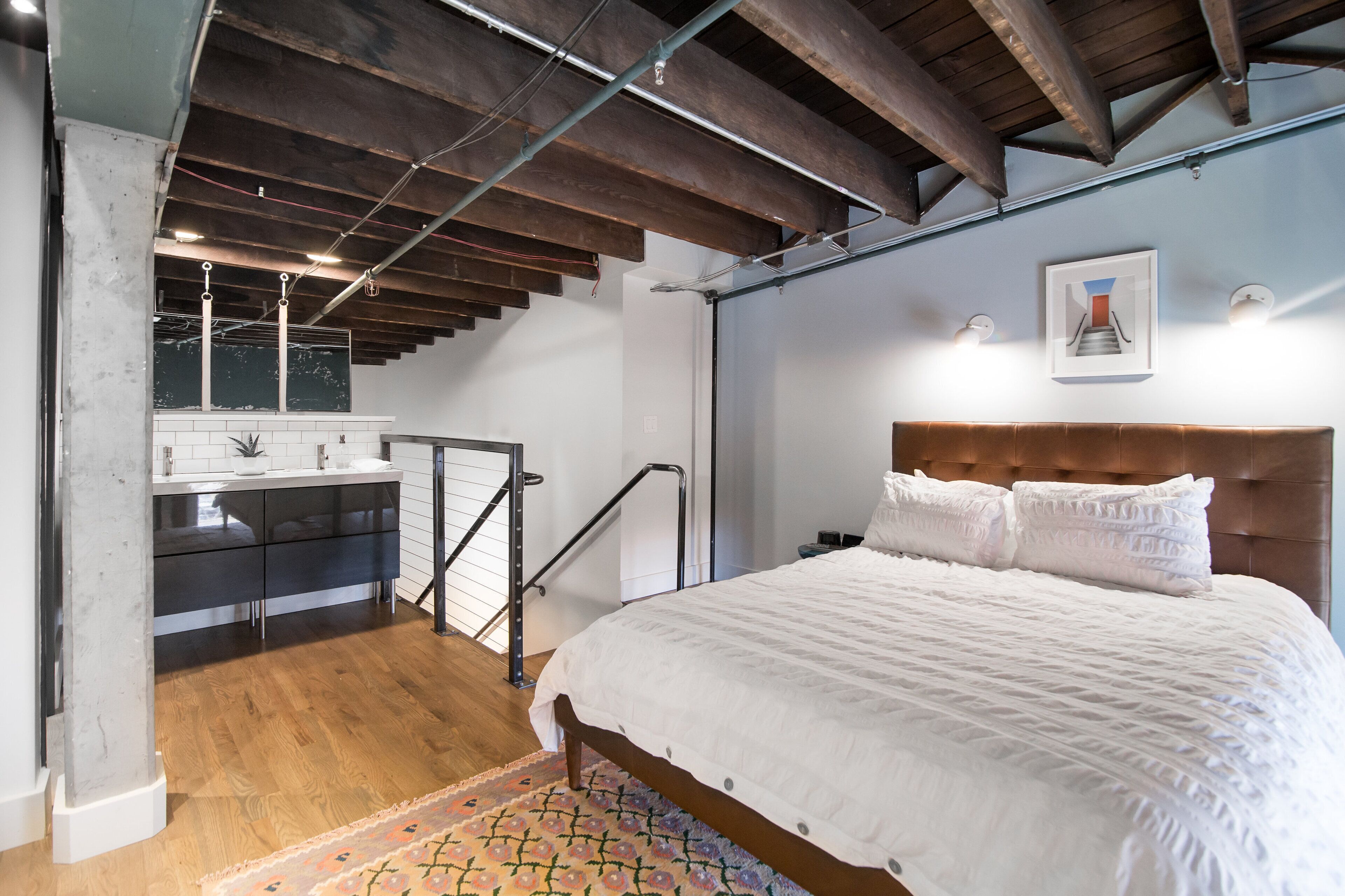 A bedroom with a large bed, wooden beams, a bathroom with a sink and mirror, and a staircase.