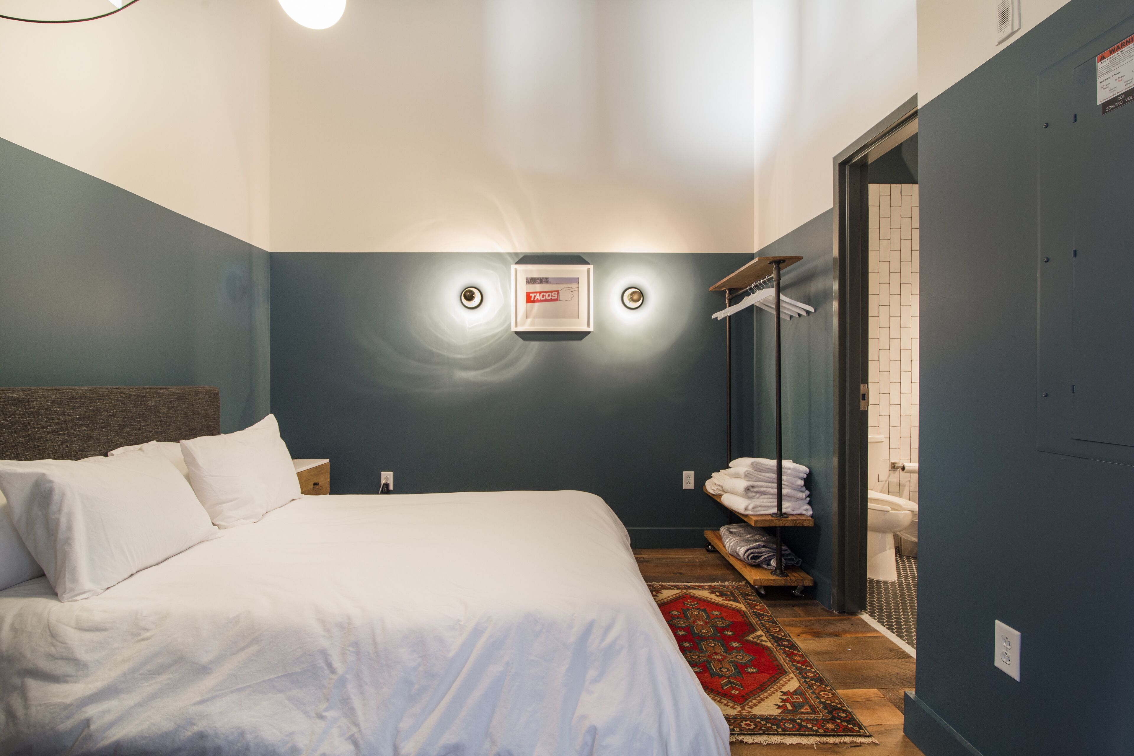 A neatly made bed with white linens, a headboard, and a small rug on the floor. There is a towel rack with folded towels and a mirror on the wall.