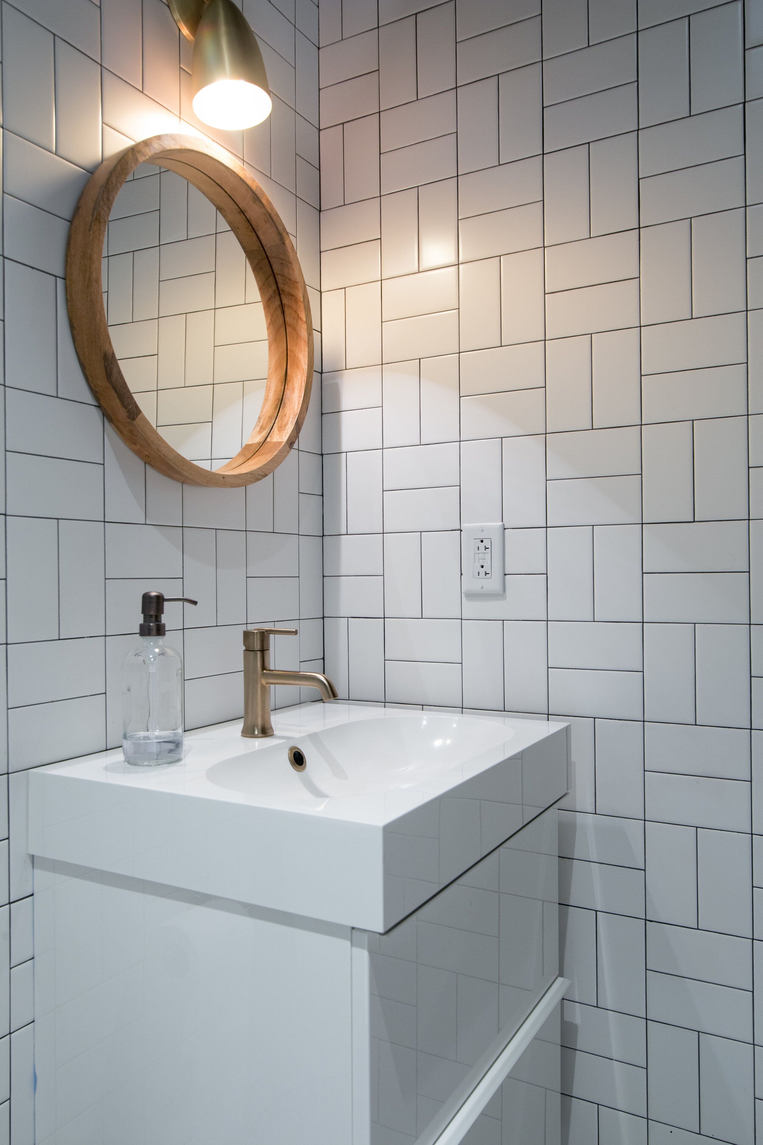 A bathroom with a white tiled wall, a wooden-framed mirror, a white sink with a gold faucet, and a clear glass soap dispenser.