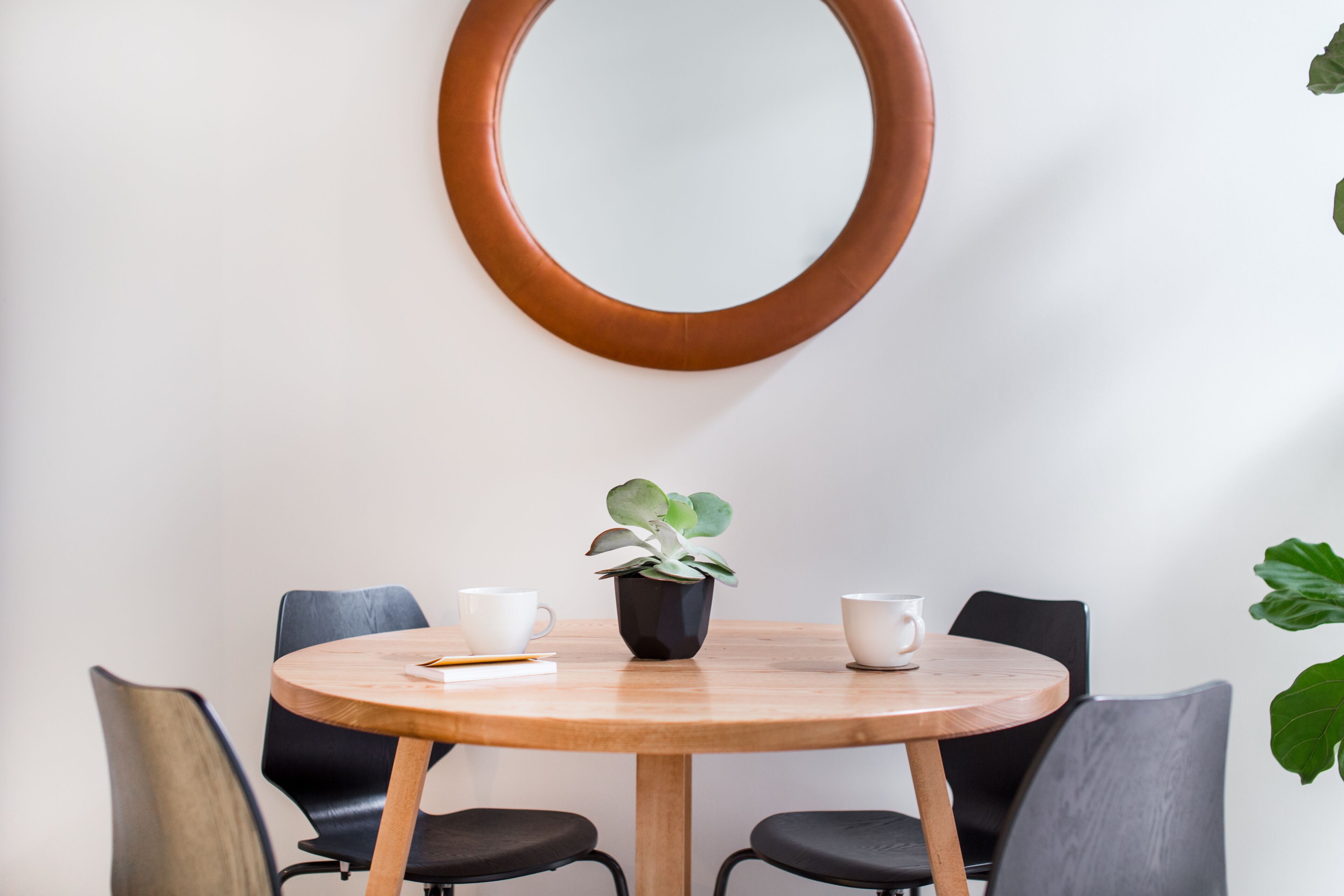 A round wooden table with four black chairs, a potted plant, and two white cups on the table.