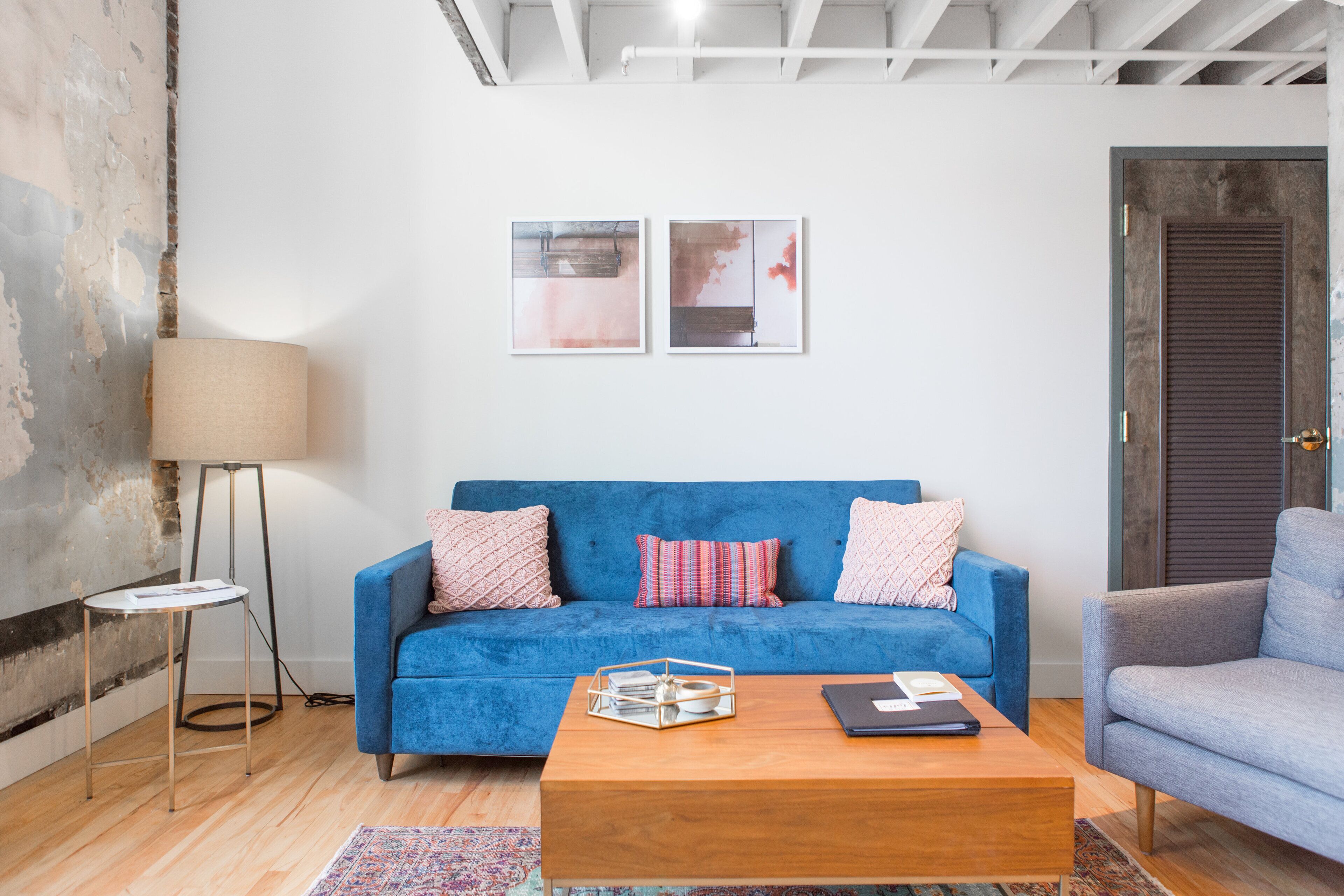 Een moderne woonkamer met een blauwe bank, een houten salontafel en een vloerlamp. Twee kunstwerken in lijsten hangen aan de muur en aan de rechterkant is een deur te zien.