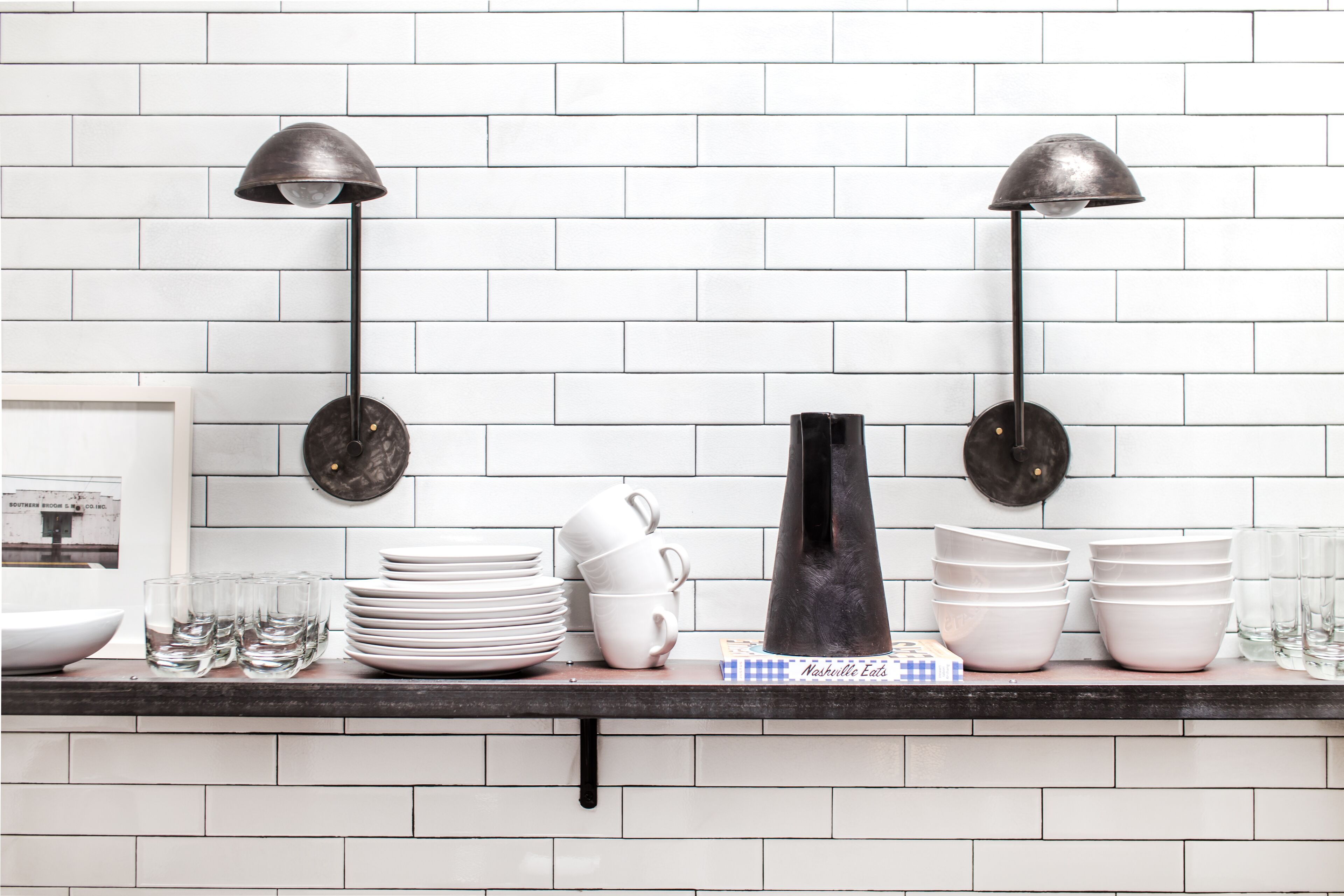 Una encimera de cocina con salpicadero de azulejos blancos, una lámpara de pie negra y diversos utensilios de cocina, incluyendo platos, tazas y un jarrón.