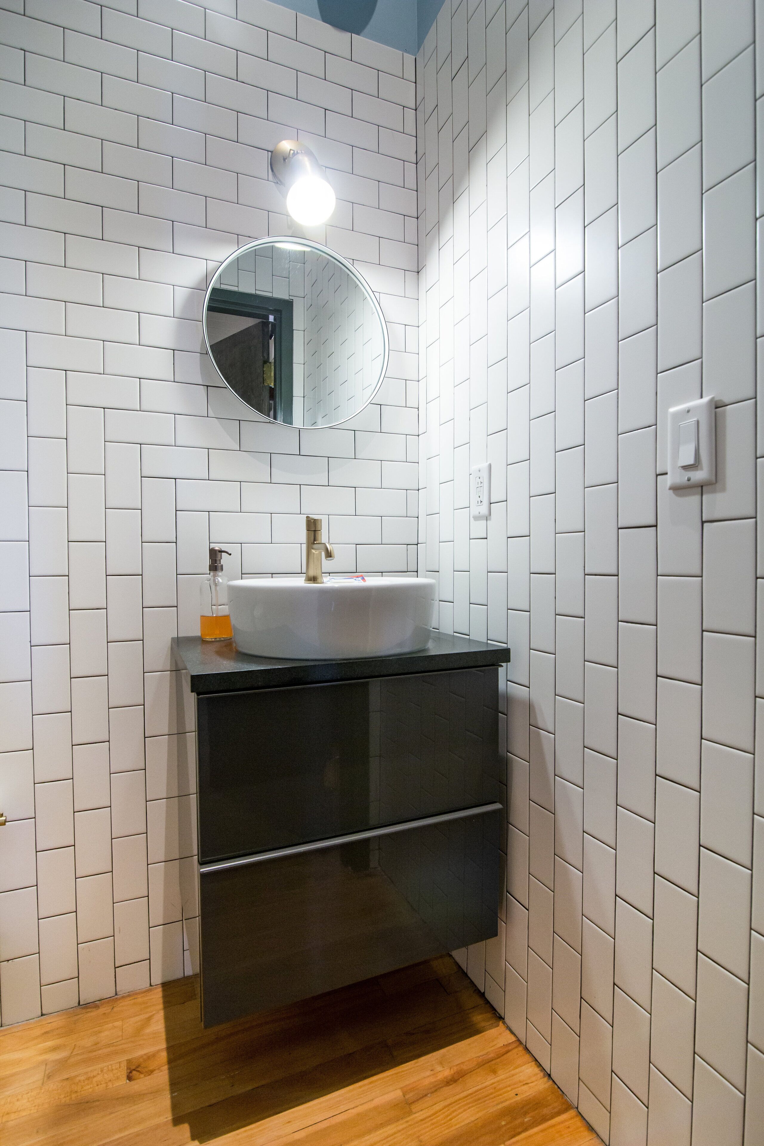 Ein Badezimmer mit weiß gefliesten Wänden, einem runden Spiegel, einem weißen Waschbecken mit goldfarbenem Wasserhahn und einem dunklen Holzschrank.