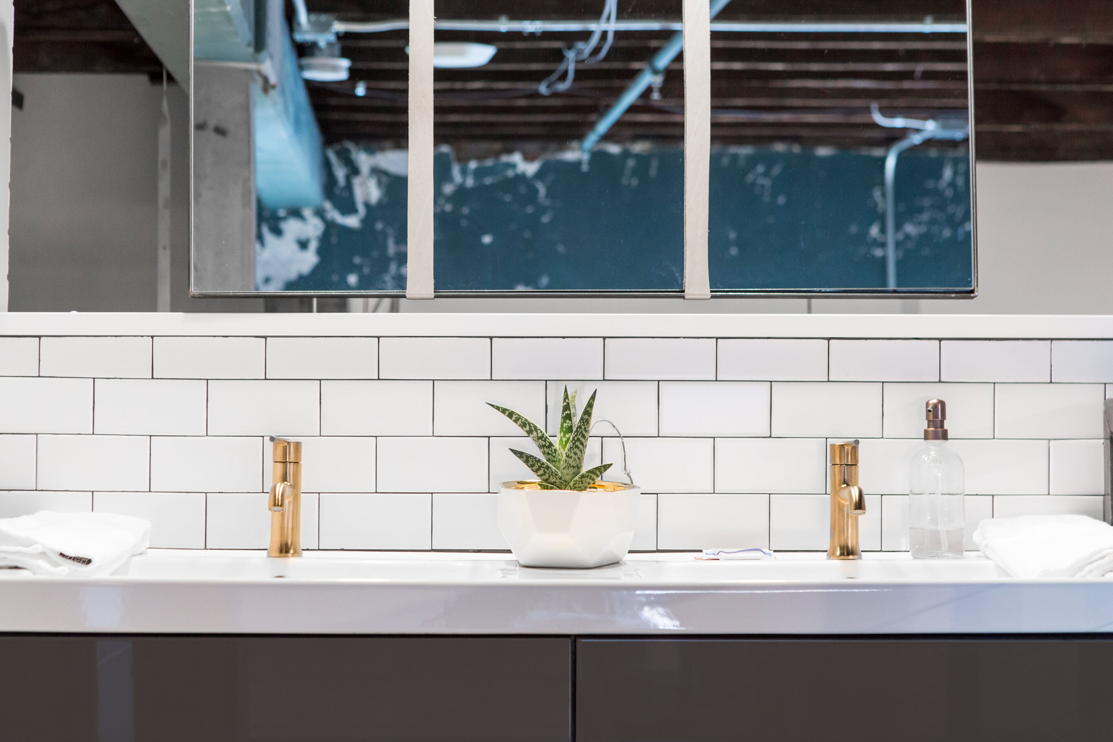 A bathroom with a white tiled backsplash, a sink with two gold faucets, a white vase with a plant, and a mirror above the sink.